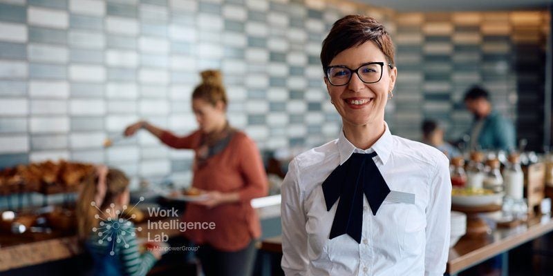 Profesional de hostelería sonriendo en un restaurante con el equipo trabajando al fondo